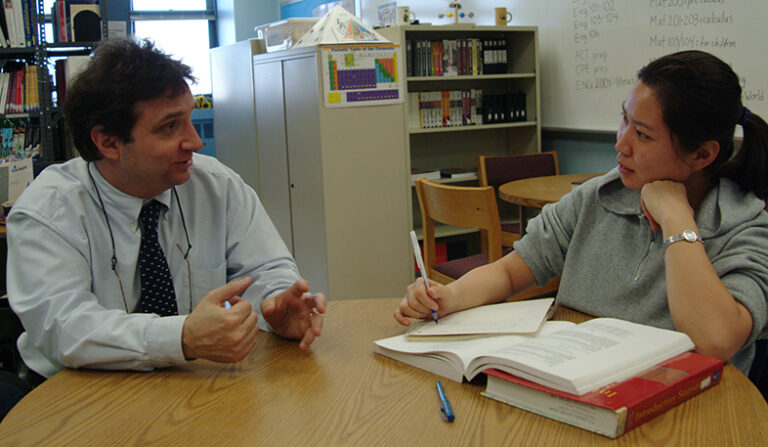 Two people having conversation while seated at a table in a classroom. Educator is on left wearing tie, student on right with notebook and textbooks.